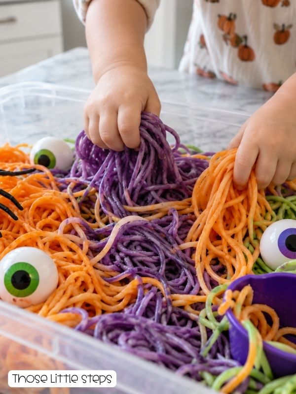 Toddler hands exploring colorful orange, purple, and green spaghetti with oversized plastic spiders and eyeballs in a shallow sensory bin.