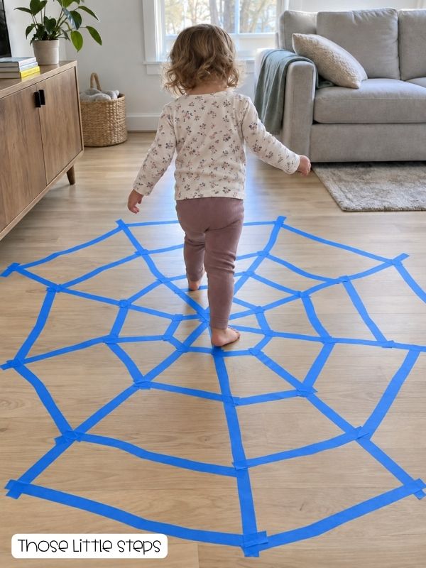 Toddler walking across an uneven painter’s-tape spider web on a living room floor for a Halloween movement activity.